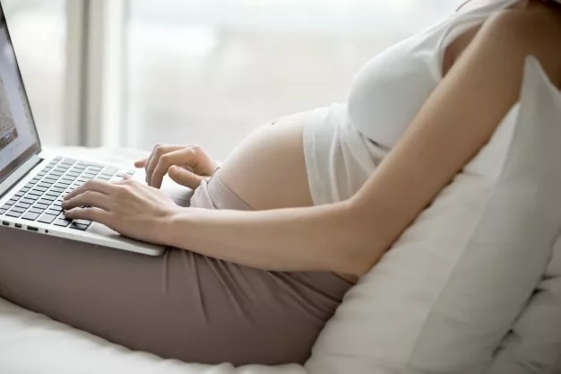 Pregnant mom sitting down on the bed and using her laptop. 